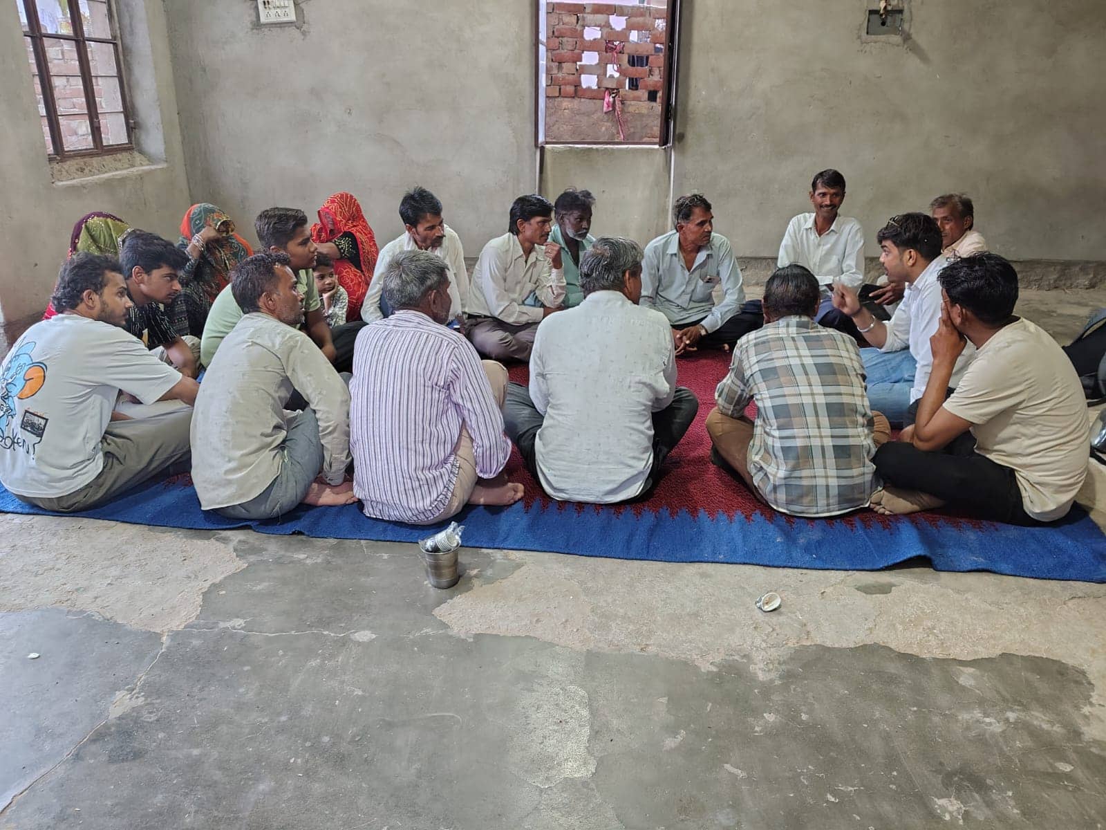 Community members, including older men and women with covered heads, sit in a circle on a red-and-blue durrie in a plain room, in discussion.
