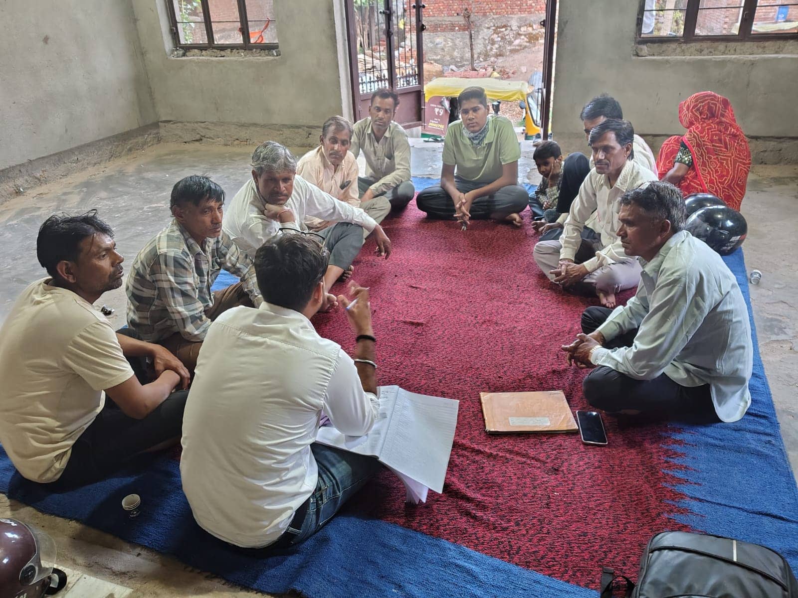 Men and a young woman sit in a circle on a red-and-blue durrie while one person takes notes in a register.