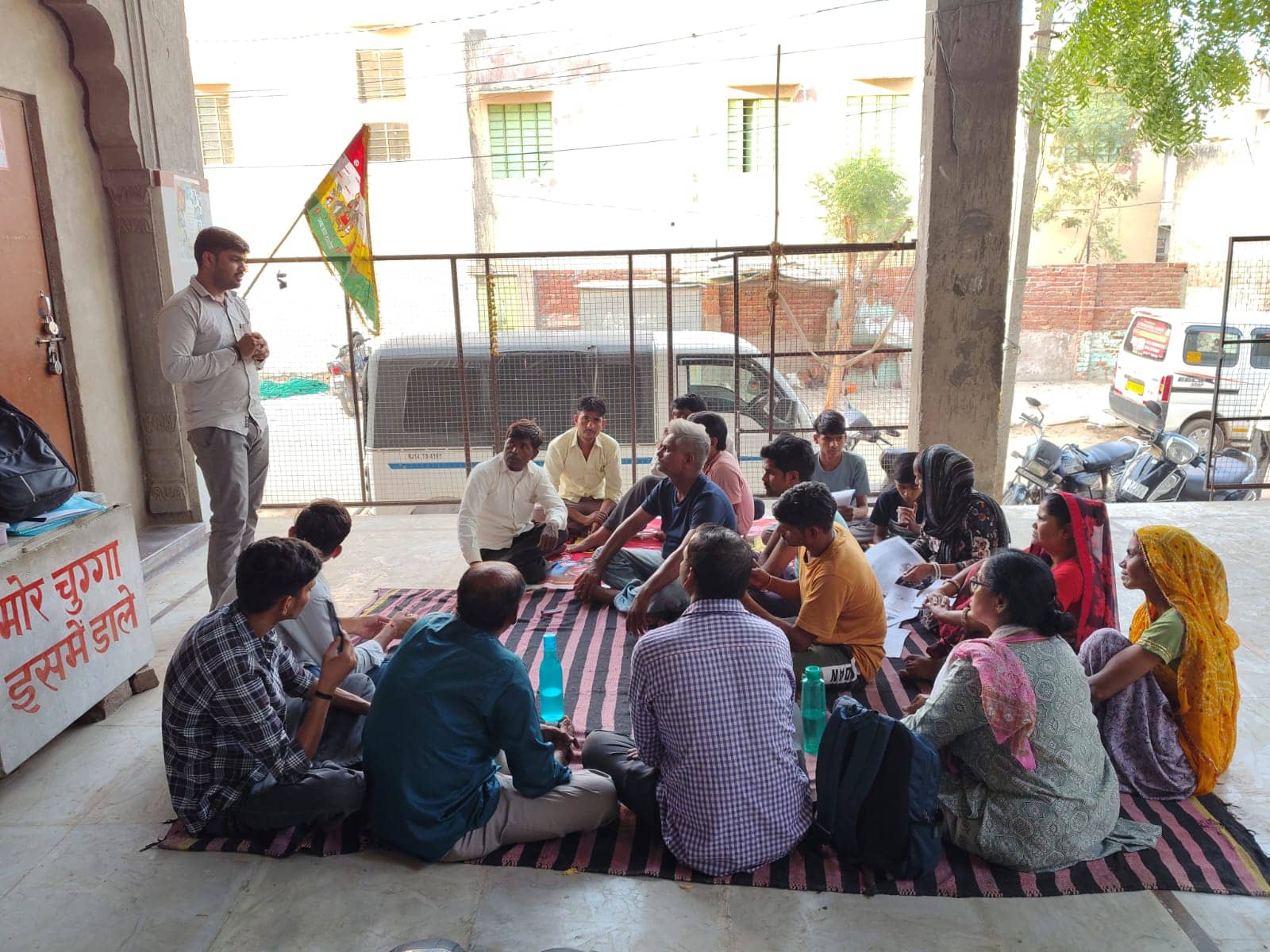 Participants seated in a courtyard listen to a speaker and look through printed sheets.