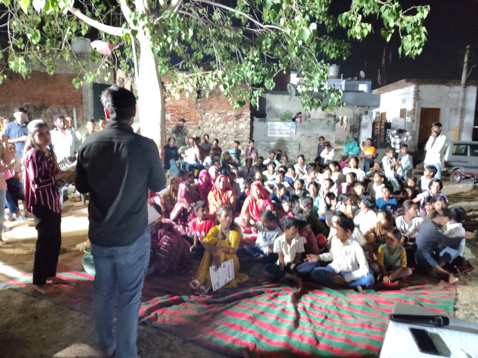 At night under a large tree, a speaker addresses women, children and men seated on a striped durrie in a neighbourhood lane.