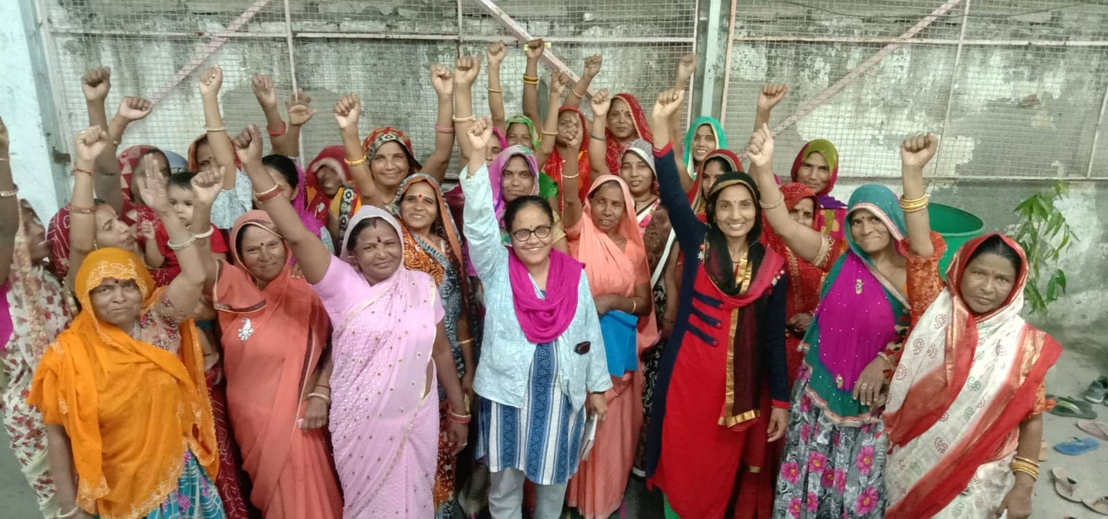 A large group of smiling women in colourful saris and odhnis raise their fists together, with a woman in a light blue kurta and pink scarf at the centre.